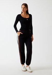 Woman standing with hands in pockets wearing a black long-sleeve top, black joggers with brown side stripes, and white sneakers.