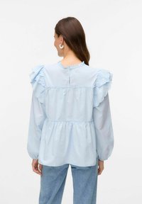Blusa azzurra chiara con spalle arricciate, vita arricciata, maniche lunghe e dettaglio di bottoni sul retro. Tessuto lucido e morbido.