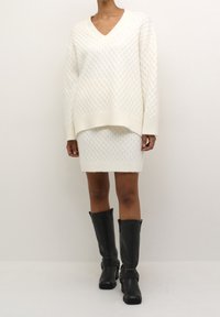 Persona che indossa un set composto da maglione lavorato a maglia bianco e gonna, con stivali in pelle neri fino al ginocchio, su uno sfondo semplice.