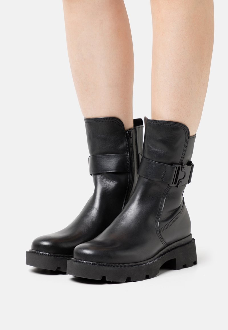 Bottes en cuir noir mi-mollet avec semelles épaisses à crampons, fermeture éclair latérale et sangle réglable à boucle portées sur des jambes nues sur fond blanc.