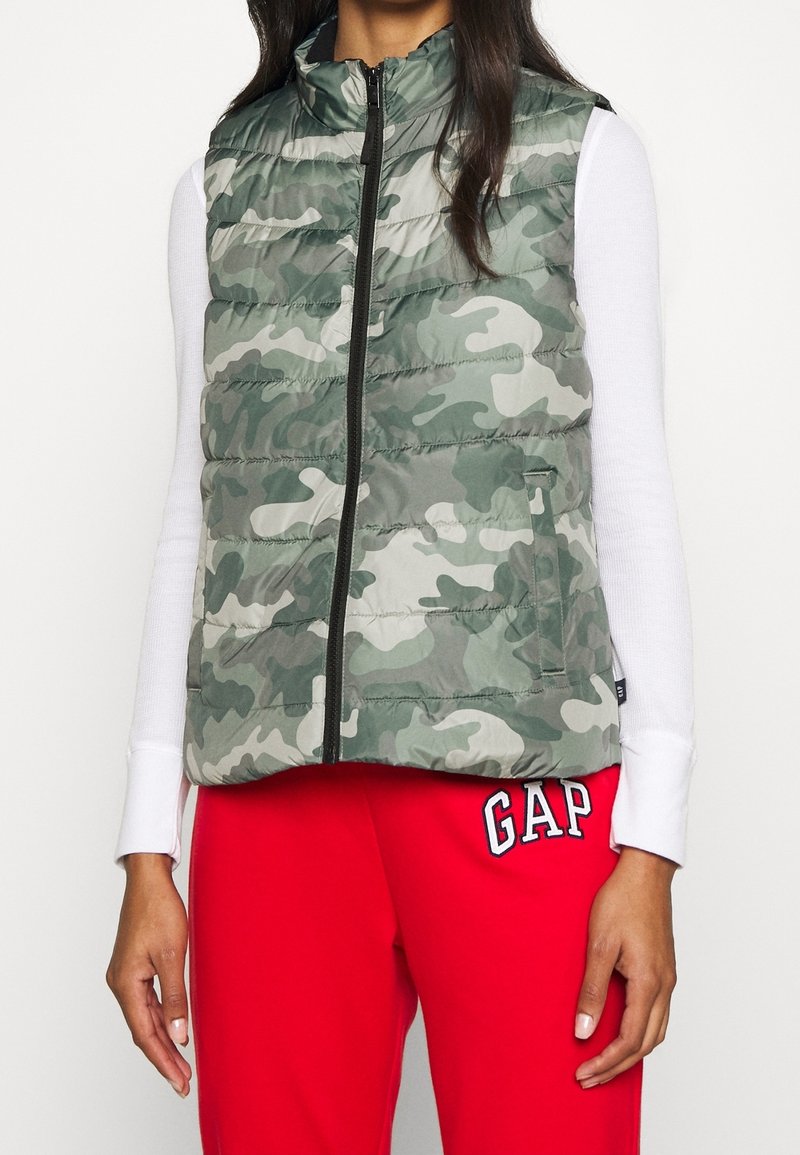 Femme portant un gilet matelassé camouflage vert et gris sur un t-shirt blanc à manches longues et un pantalon de jogging rouge avec le logo « GAP ».