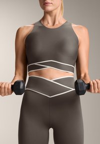 Sport crop top and high-waisted leggings in dark brown with white diagonal accents. Materials appear stretchy and fitted. User holds a dumbbell.