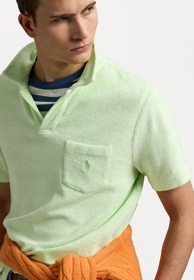 Lichtgroen polo-shirt met korte mouwen van een gestructureerde stof, voorzien van een borstzak en geborduurd logo; oranje trui om de taille geknoopt.