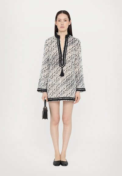 Rochie din material alb transparent cu modele abstracte monocrome, accente negre, decupaj adânc în V și detaliu cu ciucuri; asortată cu o geantă neagră.