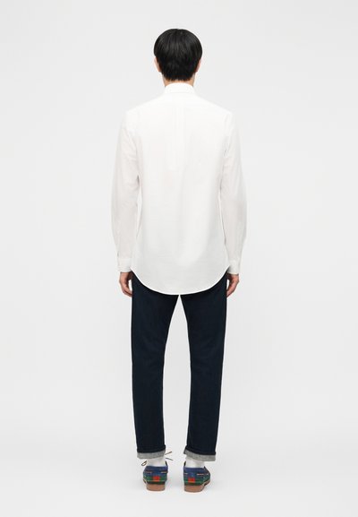 Muž oblečený v bielej košeli s dlhým rukávom, tmavé zrolované nohavice, biele ponožky a modro-hnedé topánky, stojaci chrbtom k divákovi na bielom pozadí.