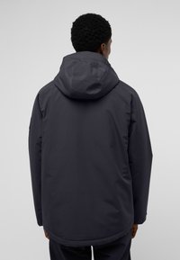 Veste à capuche gris foncé avec une texture lisse, capuche ajustable et ourlet allongé. Présente un minimum de détails et un design épuré.