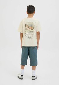 Cremefarbenes Baumwoll-T-Shirt mit einem Grafikdruck eines Autoszenenmotivs auf dem Rücken, kombiniert mit blauen Jeansshorts und schwarzen Sneakers.
