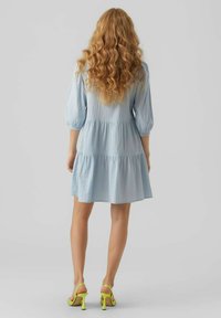 Robe bleu clair à volants avec des manches bouffantes trois-quarts, fabriquée dans un matériau léger. Portée avec des sandales à talons hauts vert fluo.