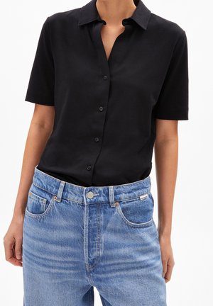 Femme portant une chemise noire à manches courtes boutonnée, rentrée dans un jean en denim bleu taille haute avec poches avant et passants pour ceinture.