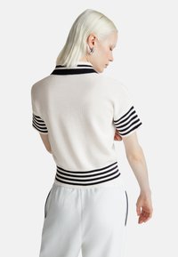 Žena s krátkými platinovými blond vlasy, oblečená v bílé pletené bluzce s krátkým rukávem, která má černé pruhované manžety a pás, v kombinaci s bílými kalhotami.