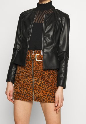 Kobieta ubrana w czarną kurtkę ze sztucznej skóry, przezroczysty czarny top i spódnicę w panterkę z zamkiem z przodu i paskiem.