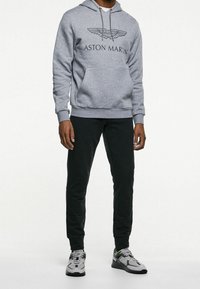 Grå hoodie med Aston Martin-logotyp, med drarem till huva och ribbade ärmslut. Matchad med svarta byxor och grå sneakers med accenter.