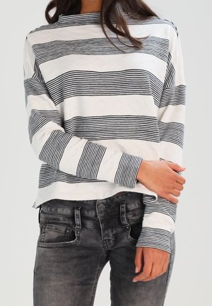 Kobieta w białym swetrze z długim rękawem w czarne poziome paski i wyblakłych czarnych dżinsach, z założonymi skrzyżowanymi na wysokości talii rękami.