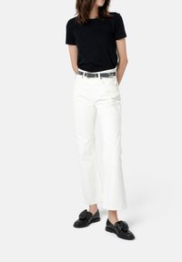 Schwarzes Baumwoll-T-Shirt, weiße weite Jeans, schwarzer Gürtel mit silberner Hardware und schwarze Schuhe mit Schleifendetail. Einheitlicher grauer Hintergrund.