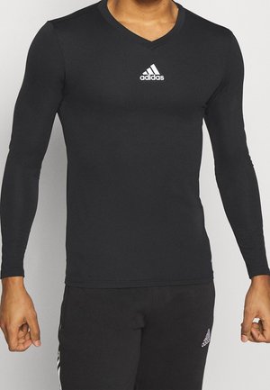 Man draagt een aansluitend zwart langemouws Adidas-shirt met logo op de borst en bijpassende zwarte Adidas-broek met logo op het dijbeen.