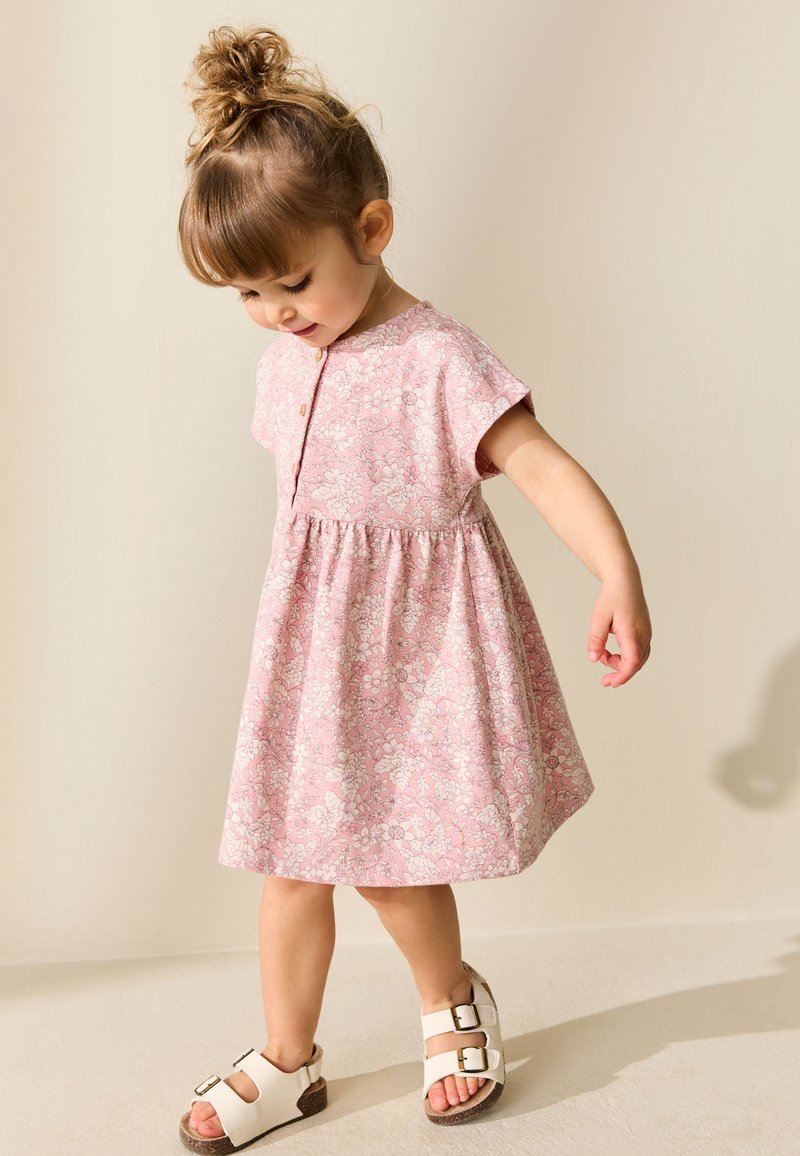 Niña pequeña con moño en el cabello, vistiendo un vestido rosa con flores y sandalias blancas, mirando hacia abajo y manteniendo el equilibrio sobre un pie.