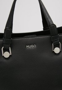 Czarna skórzana torba o fakturowanym wykończeniu, z dwoma solidnymi rączkami i srebrzystym okuciem z nazwą marki "HUGO" na środku.