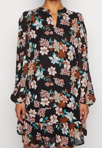 Femme portant un chemisier noir à fleurs avec des manches longues et une jupe assortie aux motifs floraux roses, turquoise et marron.
