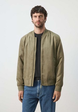 Hombre con cabello corto y rizado vestido con chaqueta verde oliva con cremallera sobre camisa negra y vaqueros azules, de pie frente a un fondo claro y liso.