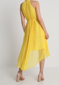 Robe plissée jaune avec un décolleté halter, taille cintrée et ourlet asymétrique, assortie à des sandales à talons nudes.