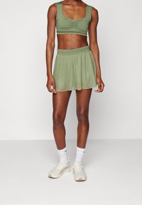 Sostén deportivo verde con fruncido y falda ligera a juego, ambos en un material suave. El modelo lleva zapatillas y calcetines blancos. Diseño sencillo.