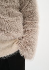 Chaqueta de piel sintética en un suave color beige con una textura larga y esponjosa. Capa inferior de punto visible en blanco roto, combinada con pantalones negros.