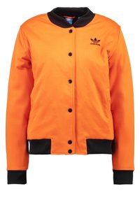 Orange jacka med svarta detaljer, knappstängning och ribbade ärmslut. Har en rundad krage och logotyp på vänster bröst. Slät textur.