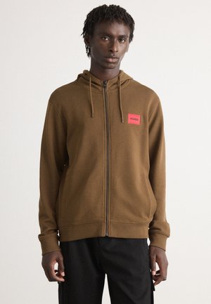 Mann in braunem Reißverschluss-Hoodie mit Kapuzenzügen und rotem "HUGO"-Patch auf der Brust, kombiniert mit schwarzen Hosen, vor einem schlichten Hintergrund stehend.
