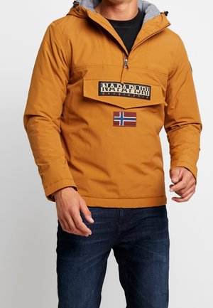Mand iført en sennepgul hættetrøje med forlommer, mærket "Napapijri geographic" og et norsk flagpatch, kombineret med mørke jeans.