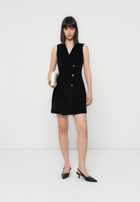 Robe noire sans manches avec un design enveloppant, comportant deux boutons, une texture lisse et un ourlet droit. Assortie à des talons aiguilles noirs.