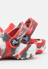 Červené, bílé a šedé vzorované nazouvací dřeváky s nastavitelnými pásky přes patu, s černým kruhovým logem s designem krokodýla.