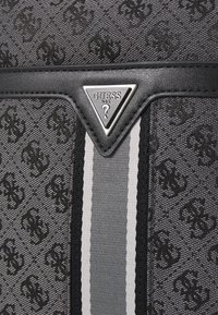 Bolso de mano negro con patrón, que presenta una placa de logo plateada, con una textura suave y dos rayas verticales grises como acento.
