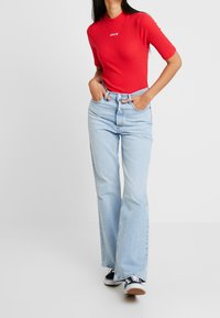 Červený žebrovaný top s krátkým rukávem a logem Levi's, kombinovaný se světle modrými zvonovými džíny a černými teniskami s bílými akcenty.
