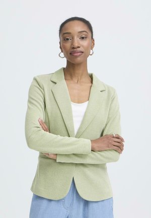 Donna sicura di sé con capelli corti, orecchini a cerchio, blazer verde, top bianco e jeans blu, in piedi con le braccia incrociate davanti a uno sfondo bianco.