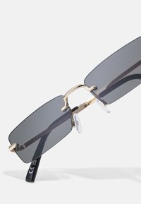 Ochelari de soare rectangulari cu lentile gri, accente de metal aurie și tampoane nazale transparente. Temele au un design elegant de culoare neagră, cu detalii ale logo-ului.