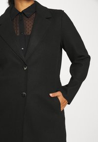 Cappotto nero con bottoni e rever indossato sopra una blusa trasparente a pois, mano in tasca, contro uno sfondo chiaro e semplice.