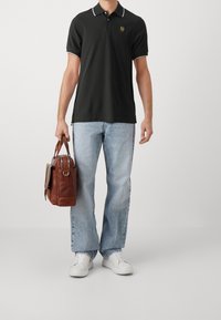 Polo noir avec bord blanc, jean décontracté bleu clair, et baskets blanches. Sac en cuir marron avec quincaillerie argentée porté à la main.