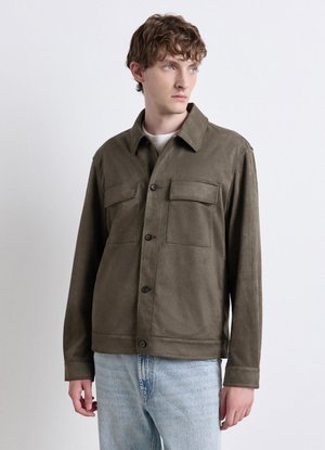 Junger Mann trägt eine braune Jacke mit Brusttaschen und hellblaue Jeans, steht vor einem schlichten weißen Hintergrund und schaut nach rechts.