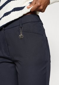 Puxar um suéter listrado em branco e azul-marinho sobre umas calças azul-marinho, com um pequeno fecho redondo prateado e preto na alça do cinto.