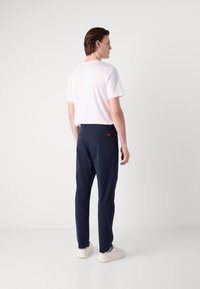 Marineblauwe chino broek met een reguliere pasvorm, voorzien van een rode label op de achterzak, gecombineerd met een eenvoudig wit T-shirt en lichtgekleurde schoenen.