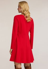 Rotes langärmliges Kleid mit tailliertem Oberteil, ausgestelltem Rock und strukturiertem Stoff. Der Rücken hat vertikale Nähte und einen runden Ausschnitt.
