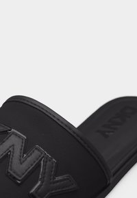 Μαύρο σανδάλι με λουρί με υφή που διαθέτει ανάγλυφα γράμματα "DKNY" και επενδυμένο πέλμα, εμφανιζόμενο σε κλειστή γωνία.