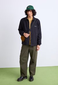 Black jacket with elastic cuffs, yellow shirt, olive green cargo pants, black platform shoes, and green cap with red logo, holding brown crossbody bag.
