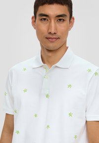 Biała koszulka polo z subtelnym haftem zielonych liści palmowych, dwoma guzikami przy plastrze oraz miękką strukturą. Casualowy design odpowiedni na lato.