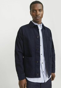 Navy blue button-up jacket in a sturdy fabric with visible stitching, two front pockets, and a collar, layered over a striped shirt.