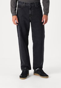 Vaqueros de mezclilla negra con un corte relajado y costuras visibles. Combinados con zapatillas deportivas negras con suela de goma. Sin patrones notables.