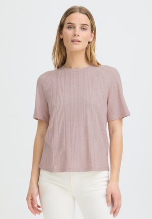 Woman with light skin and blonde hair wearing a mauve ribbed short-sleeve top and white pants, standing against a plain background.