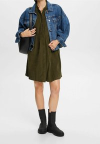 Giacca di jeans con un lavaggio azzurro chiaro, abbinata a un vestito verde oliva con vita arricciata. Stivali neri alla caviglia completano l'outfit.