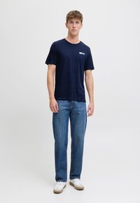 T-shirt in cotone blu navy con scollatura rotonda e maniche corte, abbinato a jeans in denim blu a vestibilità ampia e sneakers bianche.