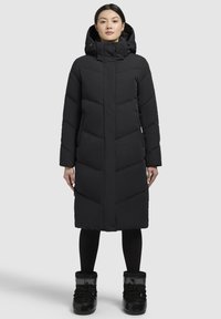 Caban lung negru cu glugă, prezentând cusături în formă de V, închideri cu capse/ butoane și buzunare laterale. Asortat cu cizme negre de zăpadă.